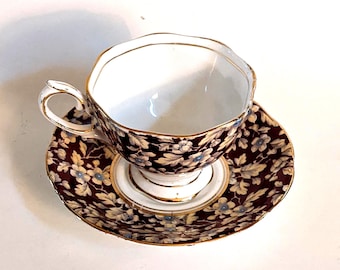 May include: A vintage teacup and saucer set. The teacup and saucer feature a dark burgundy background with a floral pattern in cream and light blue. The rims are trimmed with gold.