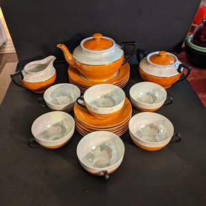 May include: A vintage tea set featuring a teapot, sugar bowl, creamer, saucers, and teacups. The set has an orange and white color scheme with black accents. The teacups and creamer have a pearlescent interior. The set includes 6 teacups.