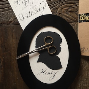 May include: A black oval frame with a white background. The frame contains a black silhouette of a person's head and shoulders. The name "Henry" is written in cursive below the silhouette. The words "Happy Birthday" are written in cursive on a white card.