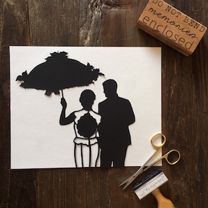 May include: A black silhouette of a couple holding an umbrella, cut out of white paper. The paper is on a wooden surface. The text "Do Not Bend memories enclosed" is on a wooden stamp.