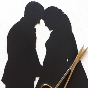 May include: A black paper silhouette of a couple, with the man in a suit and the woman in a dress, their heads touching. A pair of gold-handled scissors lies in front of the couple, suggesting a wedding or relationship theme.