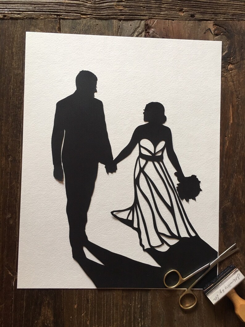 May include: Black paper cut-out silhouette of a bride and groom holding hands. The bride is wearing a gown with a detailed design. The groom is wearing a suit. The artwork is on a white background.