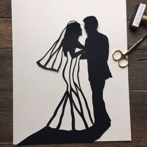 May include: Black paper cut-out silhouette of a bride and groom dancing. The bride is wearing a long gown with a veil and the groom is wearing a suit. The silhouette is on a white background.