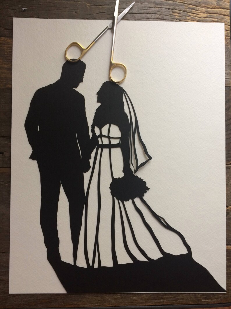 May include: A black paper silhouette of a bride and groom holding hands, with a pair of gold scissors above them.