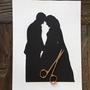 May include: A black paper silhouette of a couple in an embrace, set against a white background. The artwork is on a white paper and features a man and woman. A pair of gold-handled scissors rests on the paper.