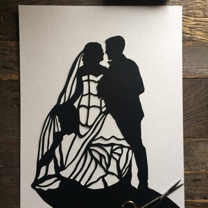 May include: Black paper cut-out silhouette of a bride and groom dancing. The bride is wearing a long, flowing gown with intricate details. The groom is wearing a tuxedo. The image is on a white background.