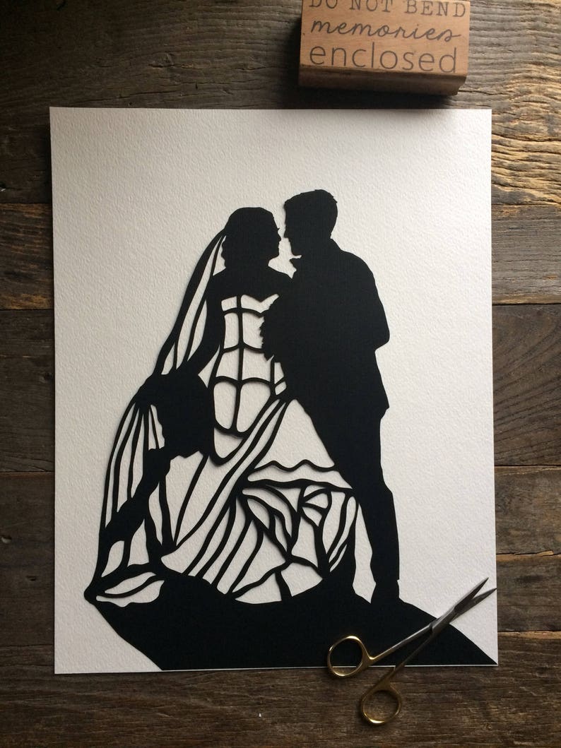 May include: Black paper cut-out silhouette of a bride and groom dancing. The bride is wearing a long, flowing gown with intricate details. The groom is wearing a tuxedo. The image is on a white background.