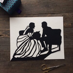 May include: Black and white paper cut-out silhouette of a bride and groom sitting on a bench. The bride is wearing a long dress with a veil and the groom is wearing a suit. The image is on a white background.