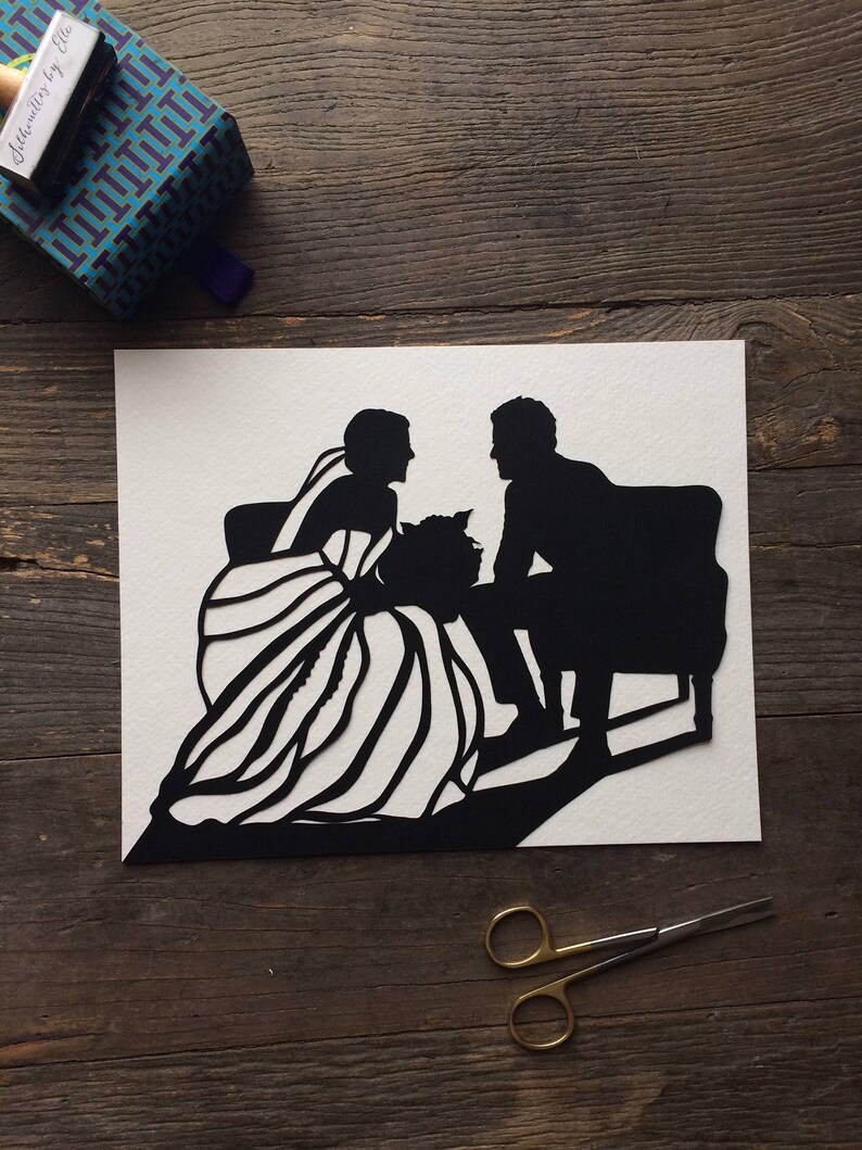 May include: Black and white paper cut-out silhouette of a bride and groom sitting on a bench. The bride is wearing a long dress with a veil and the groom is wearing a suit. The image is on a white background.
