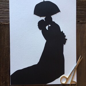 May include: A black paper silhouette of a couple kissing under an umbrella. The silhouette is cut out of white paper and is on a white background. The image is signed "Silhouettes by Ella".