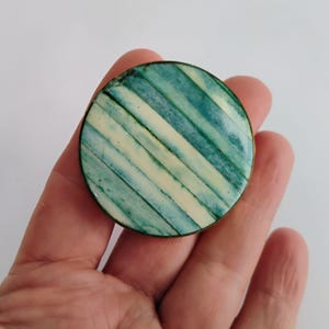May include: A round, vintage brooch with a striped pattern in shades of teal and cream. The brooch has a gold-colored metal rim and is held in a hand against a white background. The stripes are diagonal across the surface.