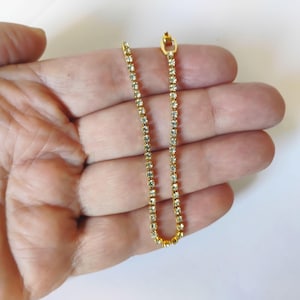 May include: A gold-tone tennis bracelet featuring a single row of clear, round-cut rhinestones set in a continuous metal chain. The bracelet includes a small, rectangular metal clasp at one end to secure the piece when worn around the wrist.