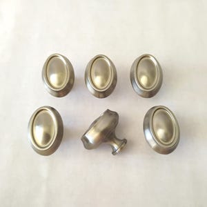 May include: Six oval-shaped cabinet knobs with a brushed nickel finish. The knobs have a raised oval center and a rounded edge. One knob is broken, showing the attachment stem.