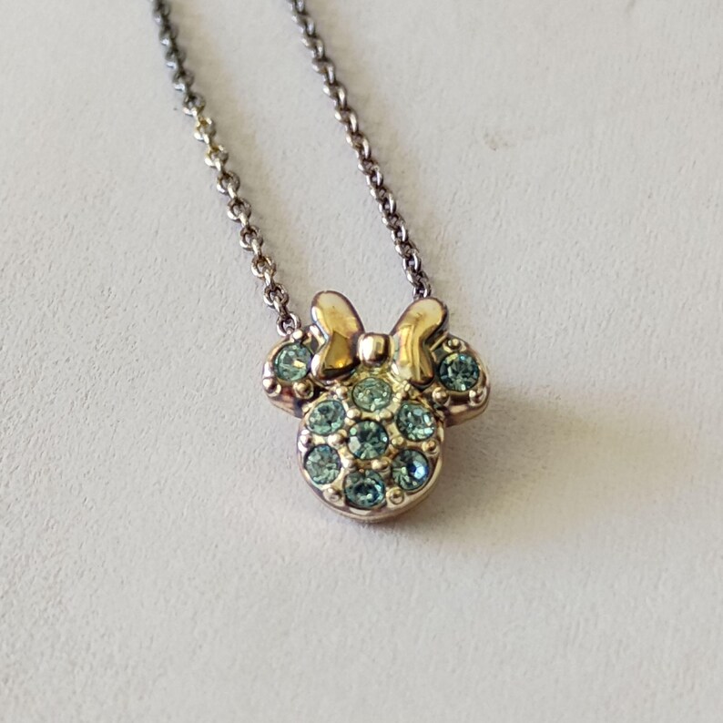 May include: A gold-toned metal necklace featuring a pendant shaped like a mouse head with a bow. The pendant is embellished with multiple small, round, light blue crystals set into the metal surface, suspended from a delicate matching chain.