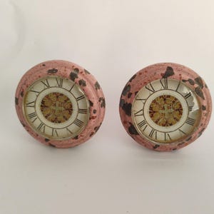 May include: Two antique-style cabinet knobs with clock faces. The knobs have a distressed pink finish with dark brown accents. The clock faces feature Roman numerals and ornate gold and white detailing, adding a vintage aesthetic.