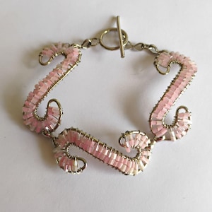 May include: A bracelet featuring three S-shaped silver-tone wire segments wrapped with small, light pink cylindrical beads. The segments are connected by silver-tone metal links and secured with a toggle clasp consisting of a ring and a bar.