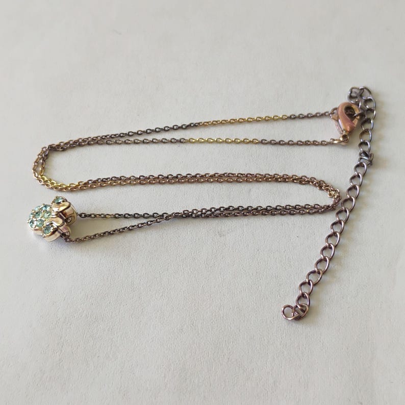 May include: A delicate necklace featuring a metallic chain with a rose gold tone and a small pendant shaped like a paw print. The pendant is encrusted with tiny, light blue crystals. The necklace includes a lobster claw clasp and an extender chain.