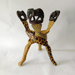 May include: A decorative wooden stand featuring three giraffe-patterned legs that intersect to support a central platform. The top of the stand is adorned with three carved dark brown elephant figures, while the central pillar has yellow and black stripes.