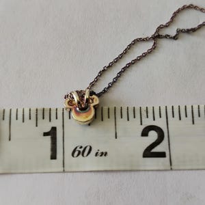 May include: A metallic pendant shaped like a stylized mouse head attached to a thin chain, positioned against a white measuring tape that shows the pendant width is approximately 0.5 inches. The pendant has a smooth, reflective surface.