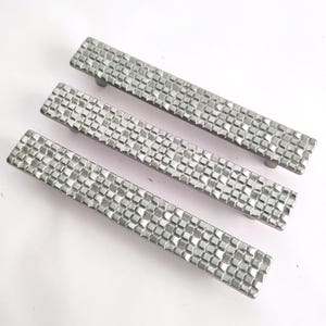 May include: Three rectangular cabinet pulls made of silver-toned metal. Each handle features a textured surface composed of a grid of small, raised square facets that create a reflective, geometric pattern across the entire length of the hardware.