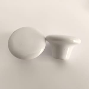 May include: Two white ceramic cabinet knobs. One knob is round, the other is a mushroom shape. Both knobs have a glossy finish and are set against a white background. These knobs are suitable for furniture restoration or DIY projects.