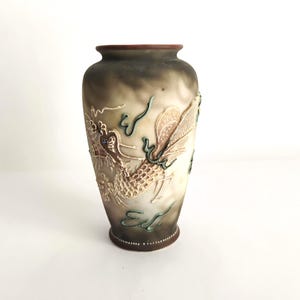 May include: A frosted glass vase featuring a raised, textured relief of a dragon with gold, white, and teal accents. The vase has a dark, smoky gradient finish that transitions from a deep brown rim to a lighter, translucent body.