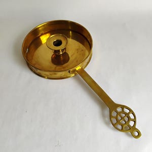 May include: A vintage brass candle holder with a circular base and a long, flat handle. The handle features a decorative, openwork design at the end. The candle holder has a central spike for securing a candle.