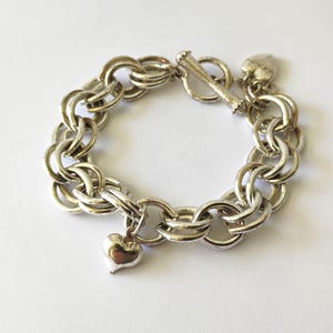 May include: A silver-toned chain link bracelet featuring a toggle clasp closure. The bracelet is constructed from double circular links and includes a small, smooth heart-shaped charm dangling from one of the links, along with a heart-shaped tag.