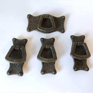 May include: Four antique brass-toned metal drawer pulls with a textured finish. Each pull features a rectangular backplate with rounded corners and a hinged, trapezoidal drop handle. The hardware has a dark, weathered appearance with visible rivets.