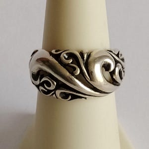 May include: A silver ring with an ornate, swirling design. The ring features a continuous pattern of flowing curves and curls, creating a textured surface. The ring is displayed on a white stand, showcasing its intricate details and craftsmanship.
