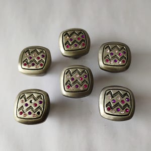 May include: Six square cabinet knobs with a silver finish. Each knob features a zigzag pattern and is embellished with pink and purple rhinestones. These decorative knobs are ideal for furniture or home decor projects.