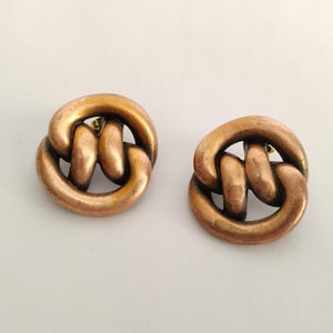 May include: A pair of circular stud earrings in a warm, burnished copper tone. Each earring features a continuous, thick metal loop that twists into an interlocking knot design, creating a rounded, sculptural shape with a smooth, metallic finish.