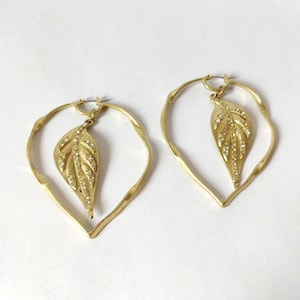 May include: A pair of gold-toned hoop earrings featuring a teardrop-shaped frame with a textured surface. Each earring includes a dangling leaf-shaped charm embellished with small, clear, sparkling crystals along the center vein and edges.