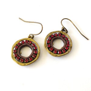 May include: A pair of dangle earrings featuring a circular brass-toned frame with an uneven, hammered texture. Each earring contains a ring of small, vibrant red crystals set against a dark brown inner circle, suspended from a curved wire hook.