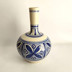 May include: A ceramic vase with a cream-colored body and a long neck. The vase is decorated with blue floral patterns and stripes. The vase has a round base and a narrow neck. The height of the vase is approximately 10 inches.