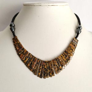 May include: A necklace featuring a central row of rectangular, tan and brown leopard-spotted stone segments arranged in a fan shape. The necklace includes small black round beads and two larger, smooth, metallic gray teardrop-shaped beads on each side.