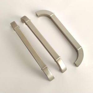 May include: Three brushed nickel cabinet pulls arranged diagonally. Each handle features a long, straight central bar with a square profile, connected to two curved end pieces that extend outward, creating a distinct architectural shape.