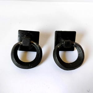 May include: Two black metal drawer pulls, each featuring a square mounting plate and a circular ring handle. The hardware has a matte finish with slight metallic wear visible along the edges of the rings, providing a consistent look for furniture.