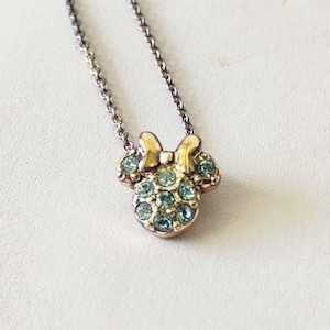 May include: A gold-toned metal necklace featuring a pendant shaped like a mouse head with a bow. The pendant is embellished with multiple small, light blue circular crystals set into the metal surface, suspended from a delicate link chain.