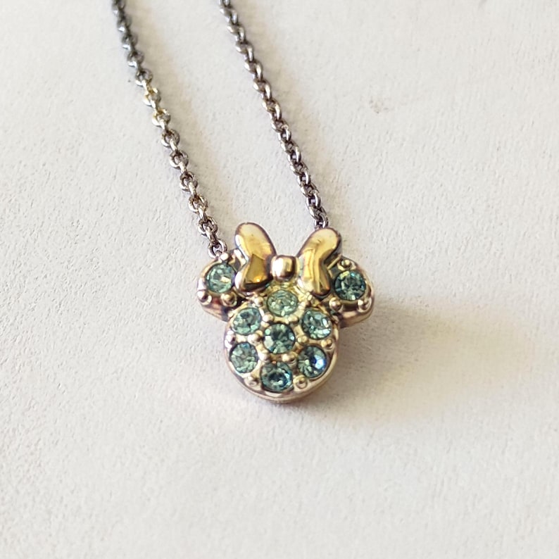 May include: A gold-toned metal necklace featuring a pendant shaped like a mouse head with a bow. The pendant is embellished with multiple small, light blue circular crystals set into the metal surface, suspended from a delicate link chain.