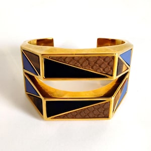 May include: Two gold-toned geometric cuff bracelets. Each bracelet features a repeating pattern of blue, black, and brown textured sections. The bracelets are displayed on a white background.