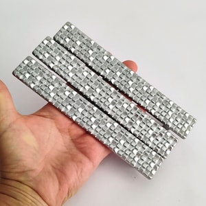 May include: Three silver rectangular bars with a textured, square pattern are held in a hand. The bars are parallel to each other and appear to be made of metal. The background is a plain white surface.