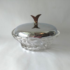 May include: A clear glass candy dish with a chrome lid and a gold-colored handle. The dish has a wavy, textured design. The lid reflects the surrounding environment. The handle is shaped like a stylized flower.