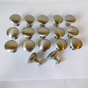 May include: A collection of seventeen silver-toned metal cabinet knobs arranged in rows. Each knob features a smooth, rounded top surface and a tapered base, exhibiting a polished metallic finish that reflects light across the circular heads.