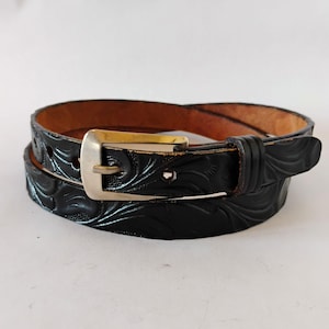 May include: A black leather belt featuring an embossed floral pattern across the strap. The belt includes a polished gold-tone metal buckle with a single prong and a matching leather keeper loop, showing a natural tan interior lining.