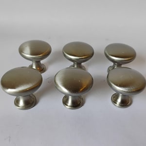 May include: A set of six round cabinet knobs with a brushed silver metallic finish. Each knob features a smooth, domed top surface and a tapered base that connects to a circular mounting plate, providing a uniform look for furniture hardware.