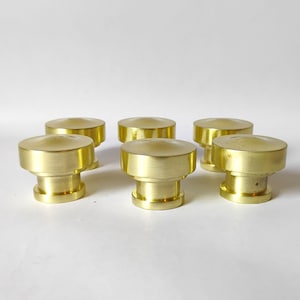 May include: A set of six circular brass knobs, each featuring a flat, slightly domed top surface and a cylindrical base. The knobs have a polished metallic finish with a warm gold tone, arranged in two rows on a plain white surface.