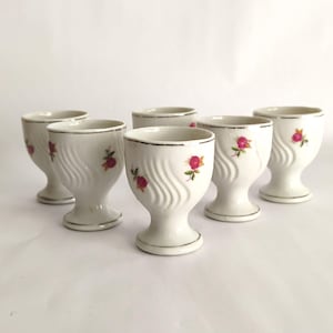 May include: A set of five white porcelain egg cups featuring a delicate embossed wave pattern and small floral motifs in pink and green. Each cup is finished with a thin silver metallic rim along the top edge and a pedestal base.
