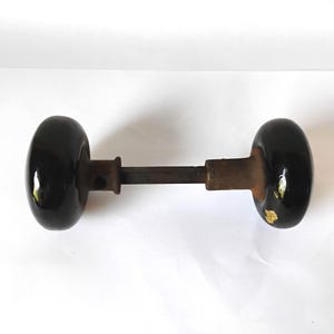 May include: A vintage door knob set featuring two round, glossy black knobs connected by a central metal spindle. The knobs have a smooth, reflective surface with minor signs of wear and oxidation on the metal components connecting the knobs.