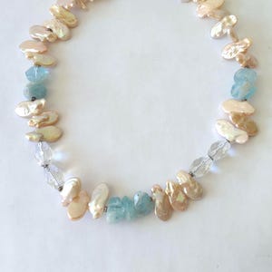May include: A necklace featuring a strand of iridescent peach-colored flat pearls interspersed with faceted light blue gemstone beads and clear, transparent crystal beads arranged in a circular pattern against a plain white background.